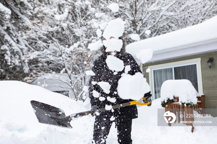 snow thrown in the air by an unrecognizable person while shoveling, snow covered house and trees in the background after a heavy snowfall.