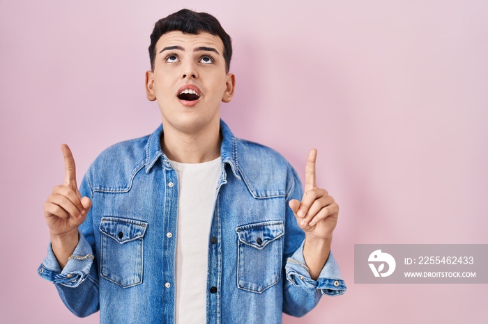 Non binary person standing over pink background amazed and surprised looking up and pointing with fingers and raised arms.