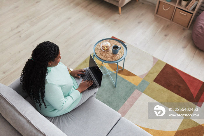 High angle view at curvy African American woman using laptop while browsing internet sitting on couch in minimal home interior, copy space