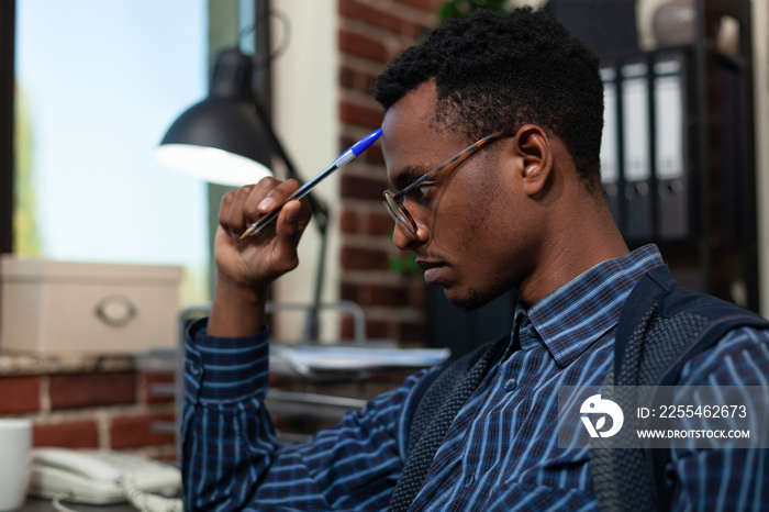 Portrait of startup employee touching forehead with pen looking focused at laptop screen with business analyitics. African american with glasses thinking about sales results sitting at desk.