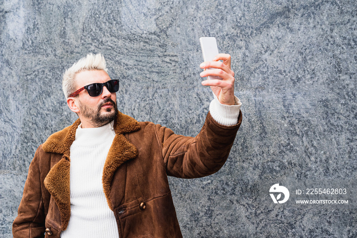 Hipster bearded influencer with platinum blonde hair and sunglasses making selfie with brand new smartphone, fashionable vlogger creating content in outdoor, copy space
