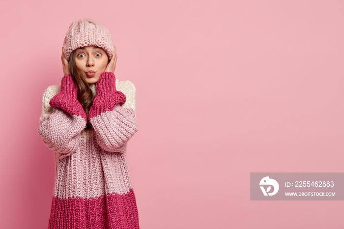 Studio portrait of young lovely woman has folded lips, makes grimace at camera wears knitted hat and loose pink jumper, isolated over rosy background with empty space for your promotion or slogan