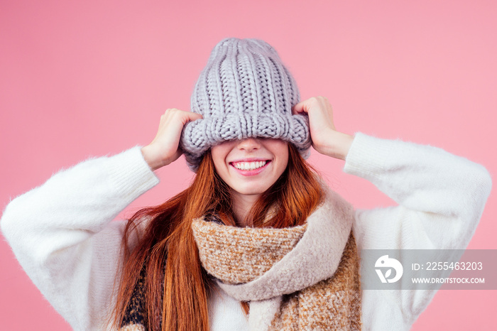 shy redhead ginger woman putting a knitted hat on his head,hiding eye and smile mouth in studio pink background