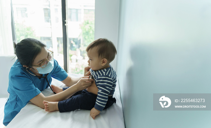 Asian Pediatrician Doctor wearing protective face mask using stethoscope to check little baby health in medical room