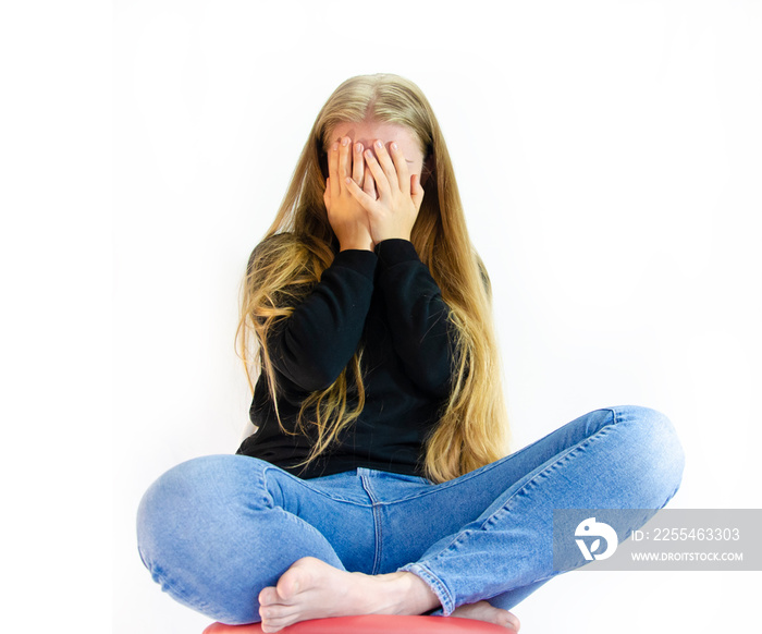 Overweight girl full legs fat woman covers her face with hands in a sitting position on a chair, dressed in simple jeans and a black sweatshirt clutching her legs on a white background