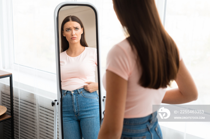 Frustrated Girl Touching Belly Standing At Mirror Slimming At Home