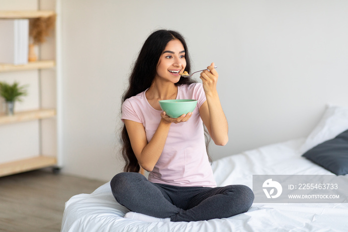 Attractive Indian lady eating yummy cereal with milk while sitting on comfortable bed at home, full length