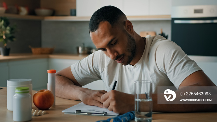 African American male nutritionist man sport fitness yoga trainer sitting in kitchen writing prescription healthy nutrition fitness diet on paper count calories with apple vitamins pills tape measure