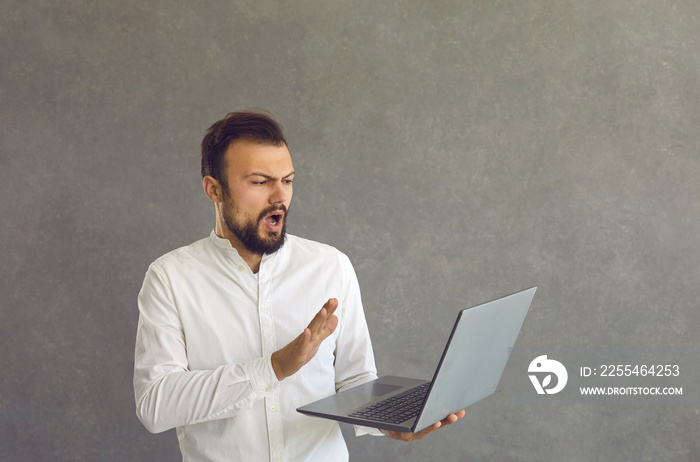 Portrait of shocked male businessman looking at laptop on gray background. Outraged puzzled Caucasian man looks at laptop screen and makes gesture of refusal saying no. Concept of negative emotions.