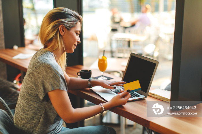 Young woman is sitting in a cafe holding a credit card with a laptop in front of her