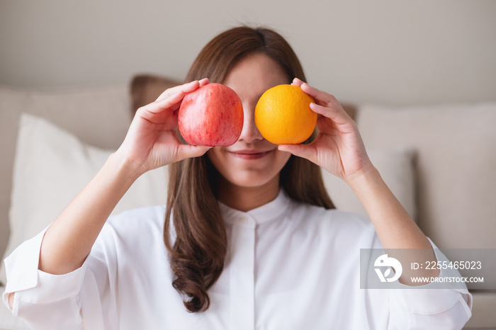 A beautiful young woman holding and covering eyes with an orange and a red apple