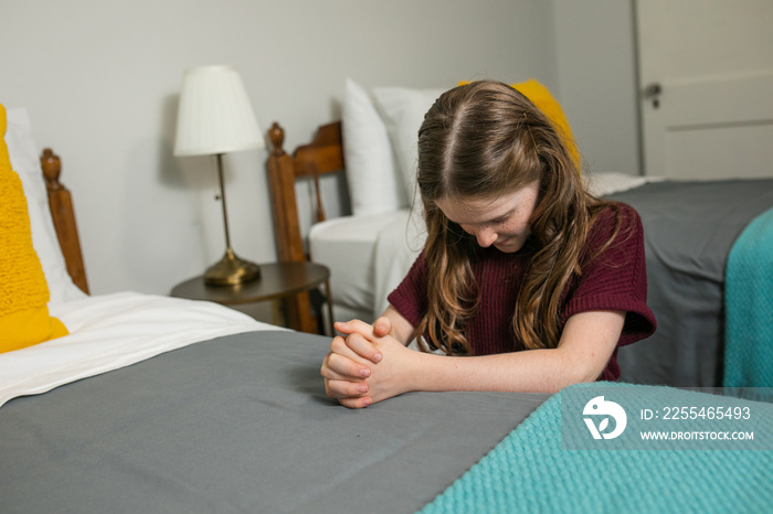 A young girl kneeling and praying to God at the side of a bed in her bedroom