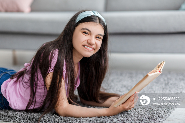 Domestic hobbies concept. Cheerful Indian teenage girl reading exciting book, lying on soft carpet at home