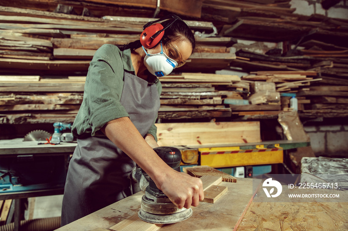 Busy and serious craftswoman grinding timbers with special machine. Beautiful woman wearing safety glasses. Concept of joiner’s shop and woodworking. Gender equality. Male profession