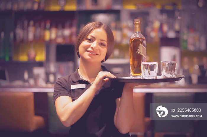 Waitress carries a whiskey glass and bottle on a tray in hotel restaurant, bar. The concept of service. Shelves with bottles of alcohol in the background. Night time.