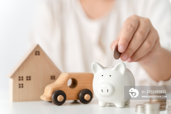 Close up of woman hand putting coin into piggy bank for saving money, stack of coins, toy house and car on table, saving money and financial concept