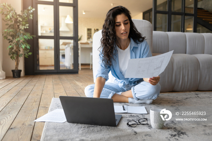 Millennial brunette woman sitting on floor with laptop and papers, working or studying from home, empty space