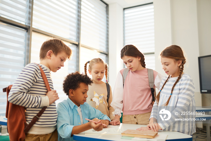 Horizontal shot of African American boy sitting at school desk doing difficult exercise, his friends standing around him ad helping with task, copy space