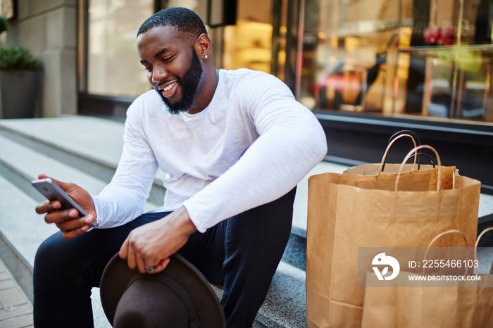 Cheerful fashion blogger writing content text for sharing publication in social media using cellphone technology during weekend shopping in city, millennial man with smartphone messaging and smiling