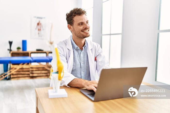 Young hispanic man wearing physiotherapist uniform using laptop working at clinic