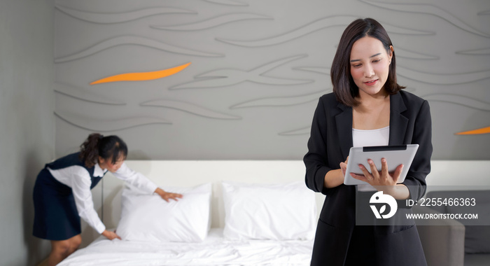 Asian businesswoman in black suit standing with tablet computer in her hand. The hotel manager verify the tidiness of the room for hotel guests while the maid making the bed in the background.