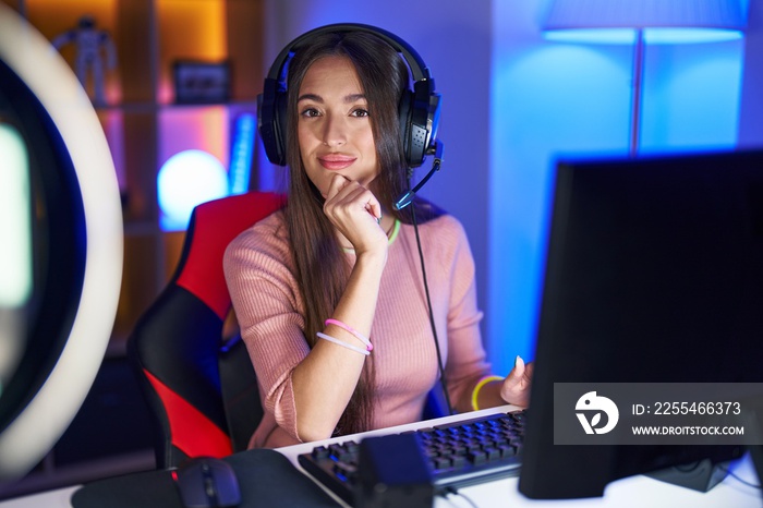 Young hispanic woman playing video games looking confident at the camera with smile with crossed arms and hand raised on chin. thinking positive.