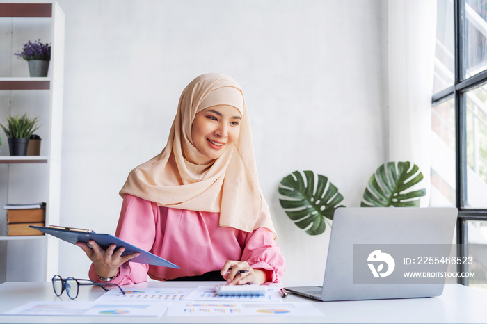 Young Asian pretty muslim lady in hijab working with laptop in office workplace, smiling islamic woman sitting at table.