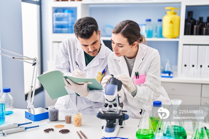 Man and woman wearing scientist uniform using microscope writing on notebook at laboratory