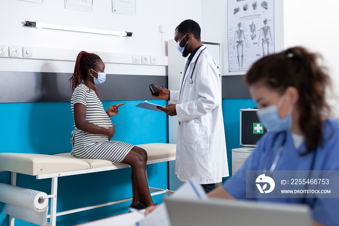 Doctor giving bottle of pills to pregnant woman at consultation. Pregnant patient talking to specialist at checkup visit, expecting child during pandemic. People wearing face masks