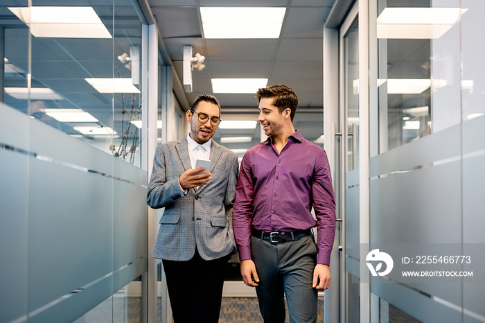 Young businessmen using mobile phone while walking through office hallway.