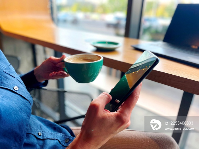 Caucasian female holding in hands cup of coffee and mobile phone having coffee break in restaurant. Morning coffee. Woman sitting in cafeteria with cellphone. Close up concept