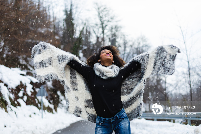 Blissful black woman raising arms under the snow in winter at mountain road.