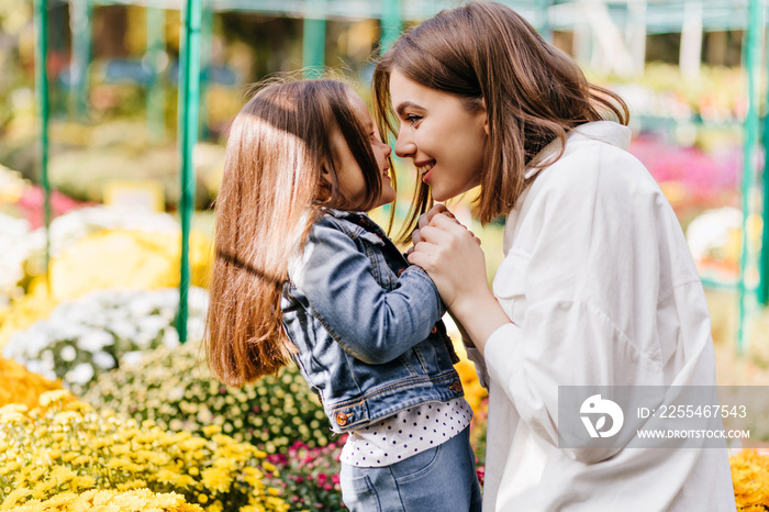 Young woman in white shirt enjoying life with child. Outdoor photo of kid and mother posing beside flowers.