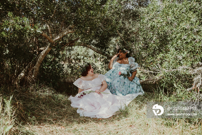 2 plus size friends wear dresses and talk while sitting under a tree