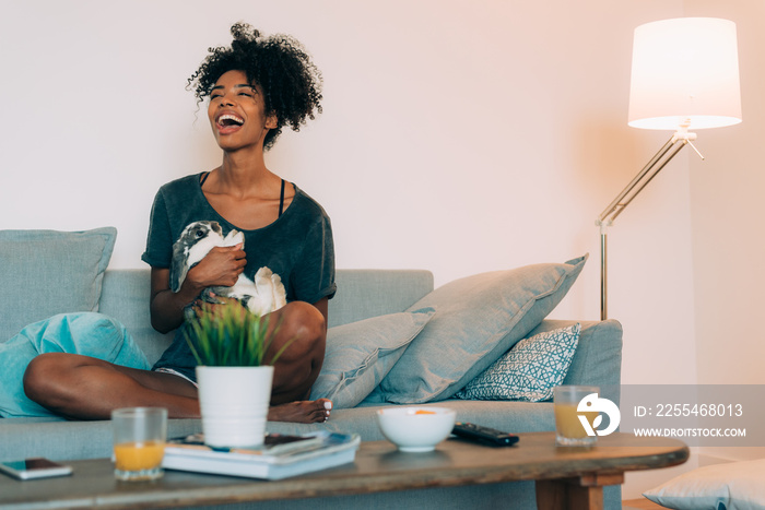 Happy young black woman sitting in the sofa giving affection to a bunny ..