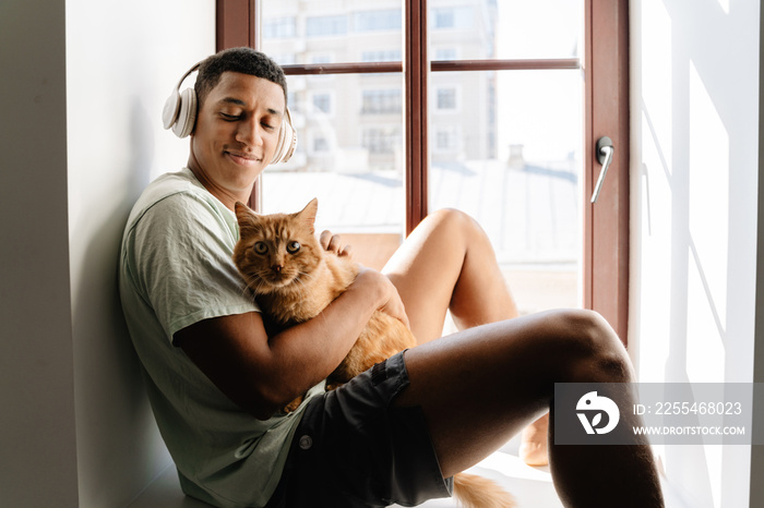 Black man in headphones sitting with his cat on windowsill