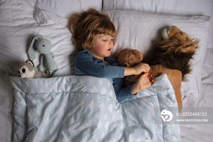 The child is getting ready for bed. Healthy child, sweetest blonde toddler boy sleeping in a bed with a teddy bear and another stuffed animals. Time before a sleeping. View from above.