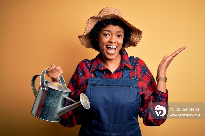 Young African American farmer woman with curly hair wearing apron and hat using water can very happy and excited, winner expression celebrating victory screaming with big smile and raised hands