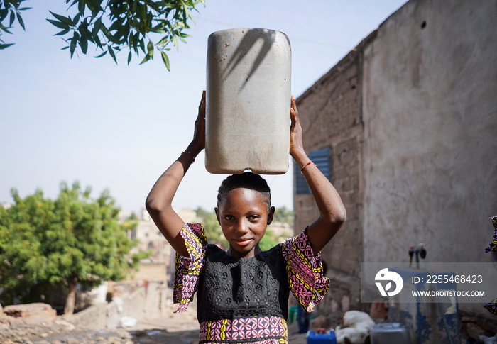 Beautiful black African village girl balancing a large canister full of water on her head that she collected from the public tap.