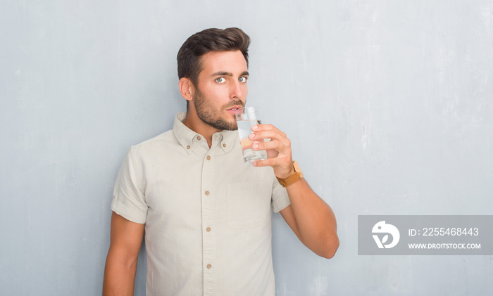 Handsome young man over grey grunge wall drinking glass of water with a confident expression on smart face thinking serious