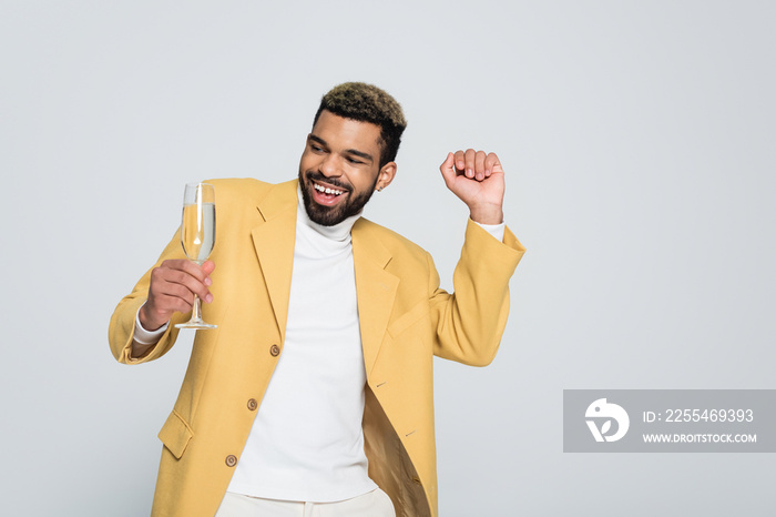 joyful african american man in stylish outfit holding glass of champagne isolated on grey.