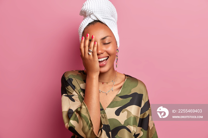 Home style concept. Joyful young woman giggles positively, makes face palm, has healthy clean skin after taking shower and cosmetic procedures, dressed in robe, poses against pink background