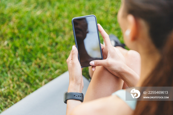 Image from back of sporty european woman 20s in sportswear resting in green park, and using cell phone with bluetooth earpod