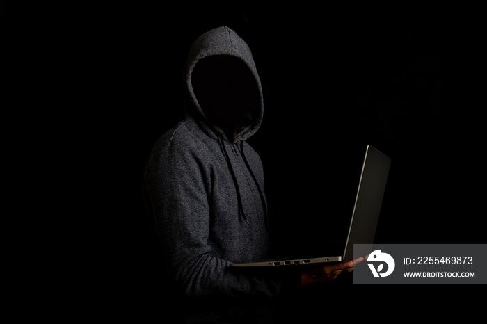 Man without a face in a hood holds a laptop on a dark background. Concept of cyber rogue, hacker. Banner