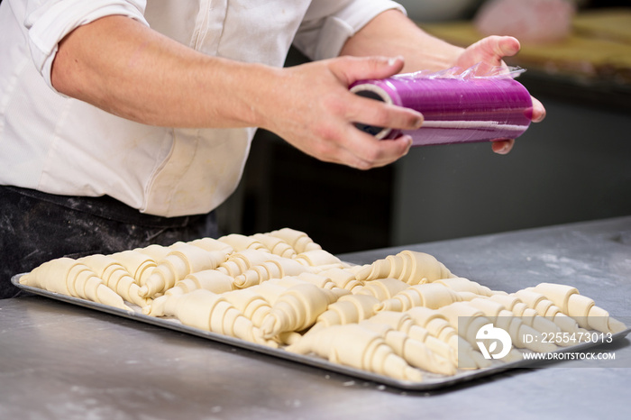 Pastry chef sealing with plastic film a tray of croissants dough .