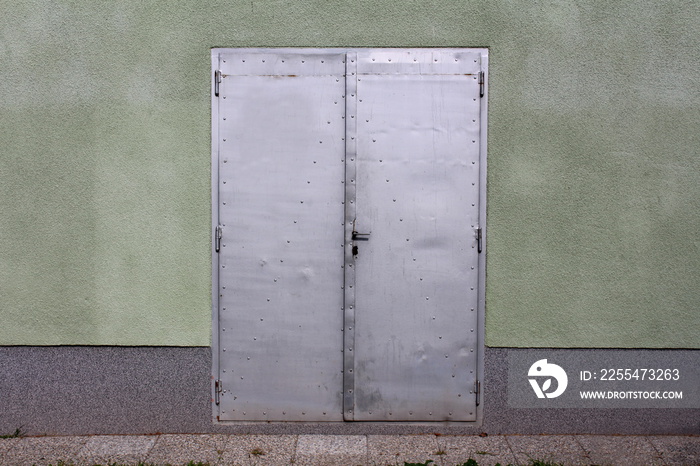 Grey sheet metal doors with small door handle mounted on house wall with stone tiles in front on warm sunny day