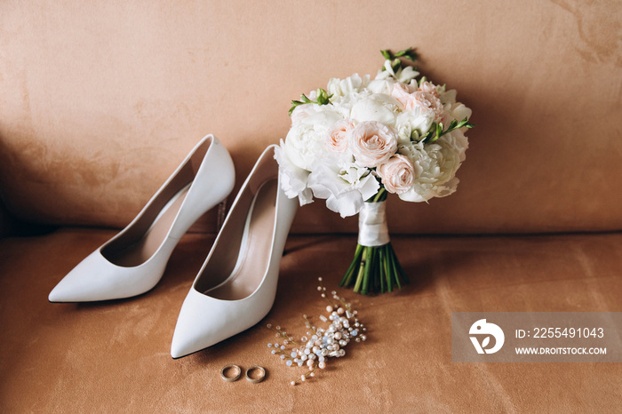 The bride’s wedding details. There are white shoes on the couch next to bouquet of white flowers and greenery with ribbon. Next to it are wedding rings and hair jewelry