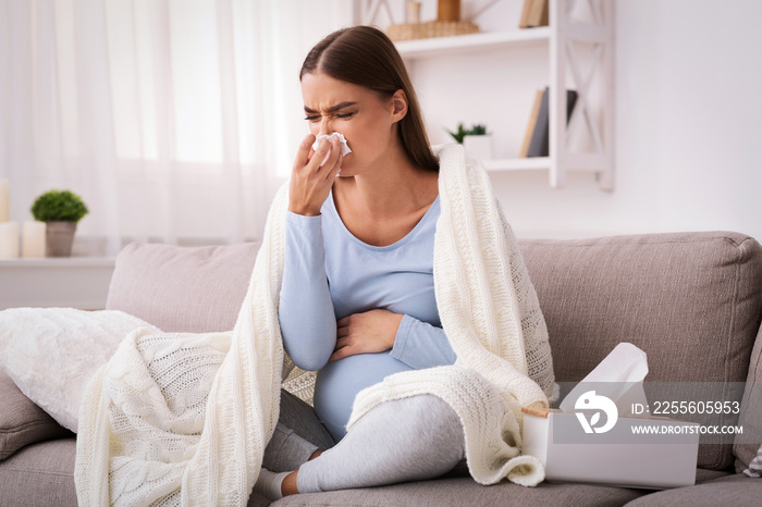 Pregnant woman blowing nose in tissue sitting on sofa indoor