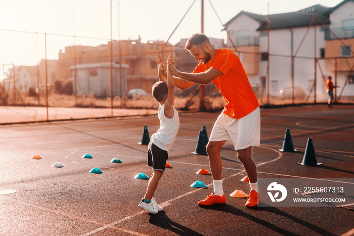 Father and son in sportswear giving high five with both hands while standing on the court in the mor