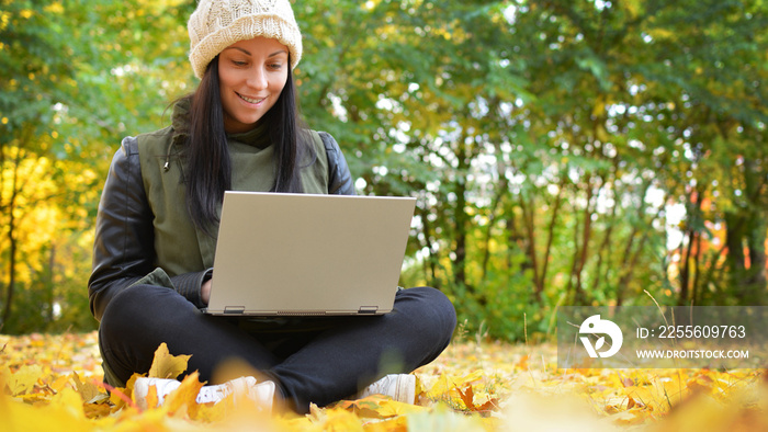 秋天公园里穿着时髦衣服拿着笔记本电脑的女孩。一个戴帽子的女人坐在地上用笔记本电脑